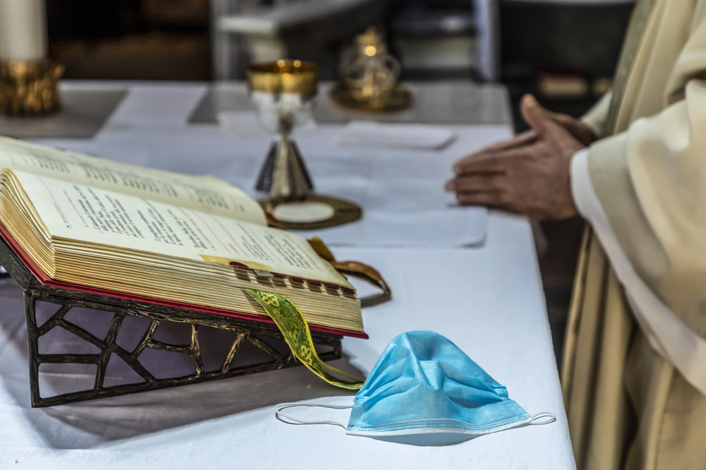 Mundschutz auf dem Altar / www.kna-bild.de / Stefano Dal Pozzolo Ein Priester faltet die Hände und feiert den Gottesdienst am Altar, auf dem das Messbuch aufgeschlagen liegt, daneben eine einfache Atemschutzmaske, dahinter liturgische Geräte, am 8. Mai 2020 in Rom.